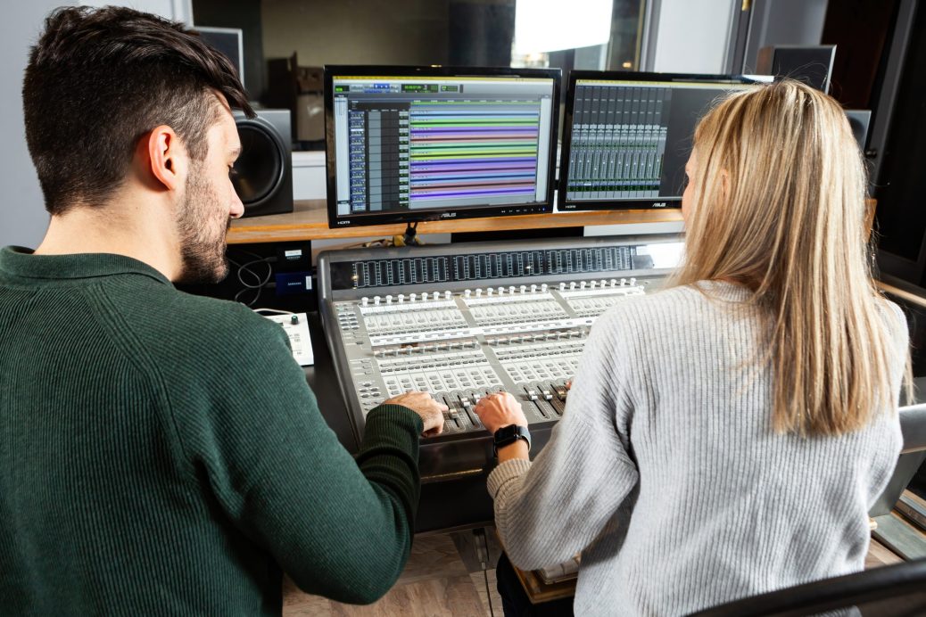 A man with black hair and a woman with blonde hair sit beside each other working on an audio board in a studio.