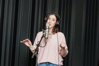 A woman wearing a pink shirt standing in front of a microphone moves her hands as she records a voice over.