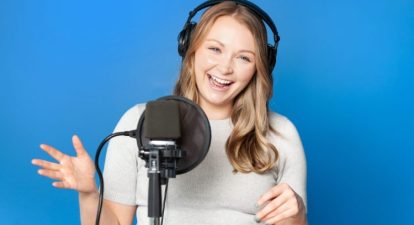 A blonde-haired female voice actor wearing headphones, smiling and speaking into a microphone.