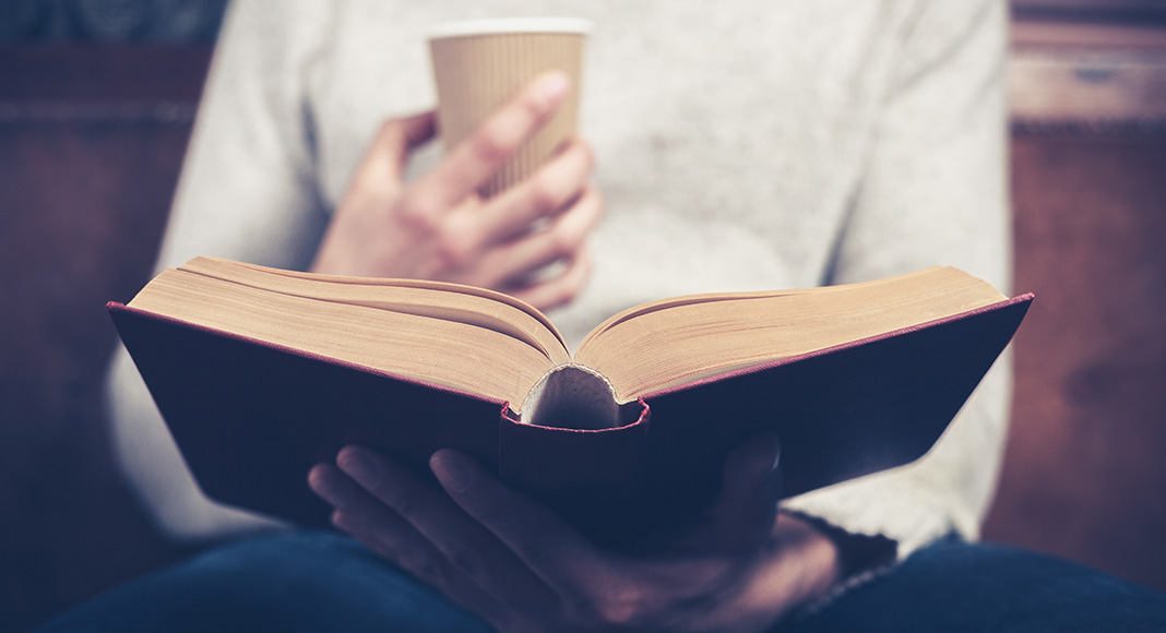 A person sits with a coffee in one hand and an open book in their lap