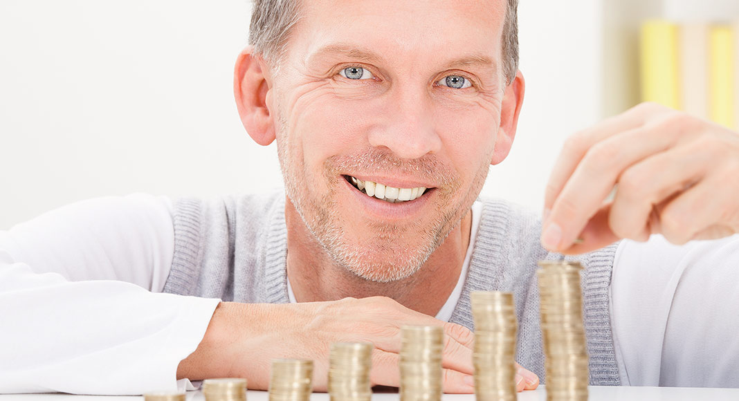 A man with blue eyes and gray hair smiles and looks at the camera as he stacks coins in several rows.