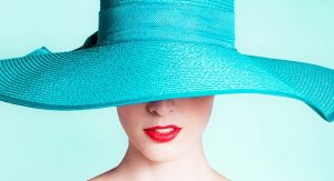 the lower portion of a woman's face is visible from underneath a high fashion hat