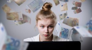 A woman looks concerned as she looks at her computer screens, while euros swirl around her