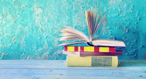 A stack of four books sits on a light blue table in front of a teal washed stucco wall