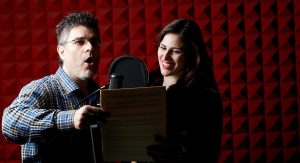 A man and a woman stand next to each other in a room with soundproofing. The man is holding a script and the woman is standing next to him, smiling. A microphone is in front of both of them.