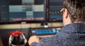 A man sits in front of a computer screen. The background is blurry, however to his left sits a set of headphones. In front of him appears to be a mixing board and vu meters. On the screen looks to be audio editing programs.