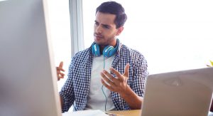 A brunette man in a plaid shirt with blue headphones around his neck stares at a computer screen with a confused expression as if he's trying to decipher a voice over script.