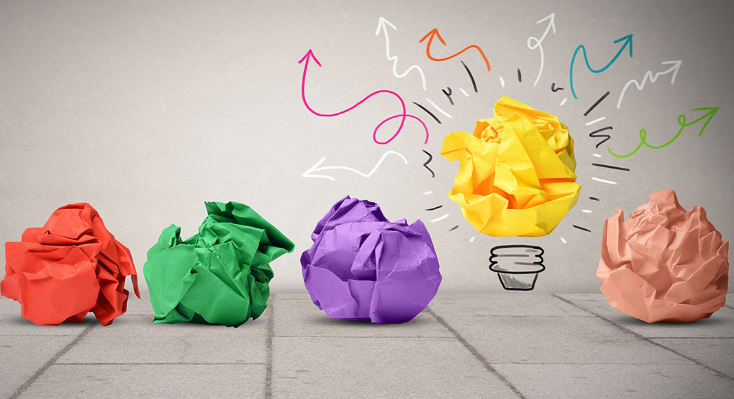 Four brightly colored crumpled pieces of paper sit on the ground while a bright yellow one is elevated and looks lit up light a lightbulb