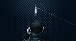 A man wearing a ballcap and headphones has his back turned to the camera as he speaks into a microphone in an isobooth