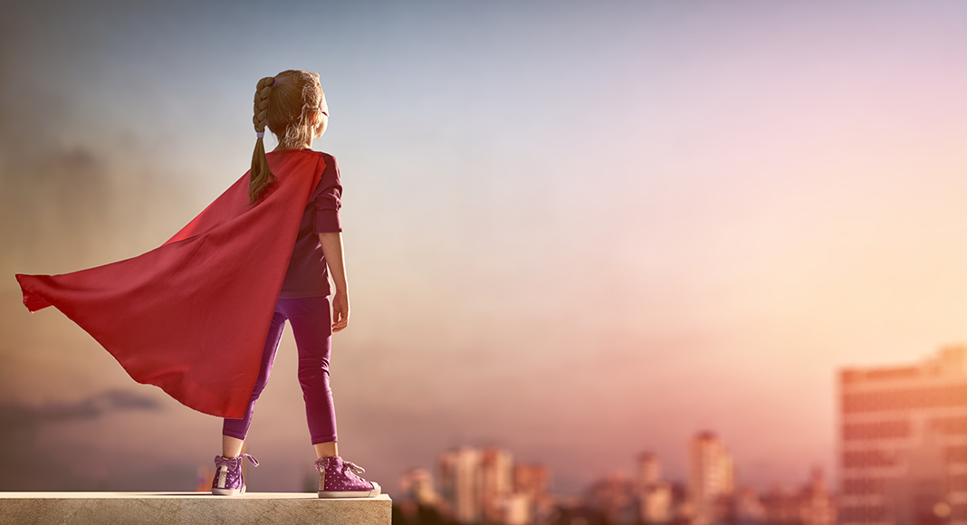 Little girl with a cape on stands at the top of a building with the wind in her hair