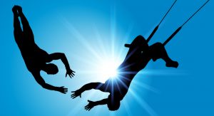 The silhouettes of two trapeze artists show one extending his hands to the other, who is in mid-air