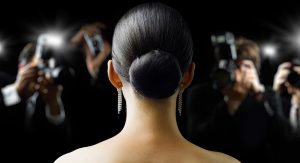View of a woman's face from behind, before her, we see that there are paparazzi taking photos. She looks like she's on a red carpet and is dressed elegantly.