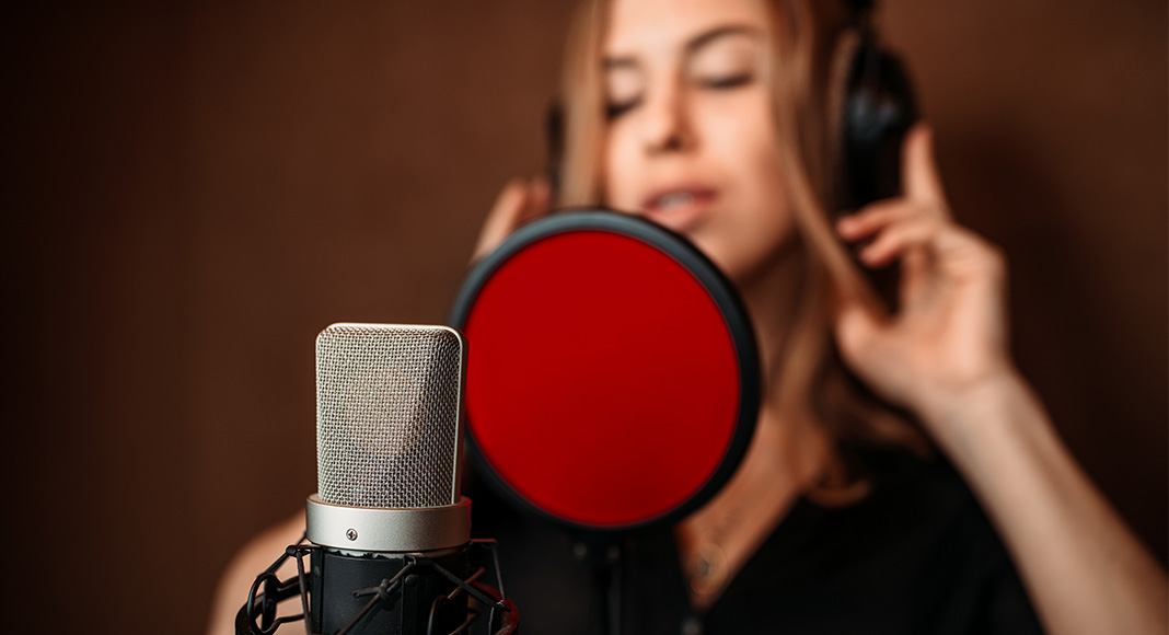 A female singer stands in the background. In the foreground is a microphone with a red pop filter in front of it.