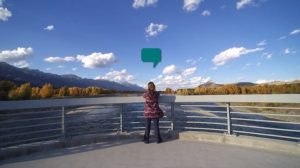 A woman stands on a bridge overlooking a river. A green speech bubble floats over her head.