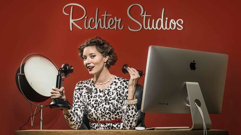 A woman sitting at a desk with a computer and a studio light, and she is speaking into a vintage candlestick phone.