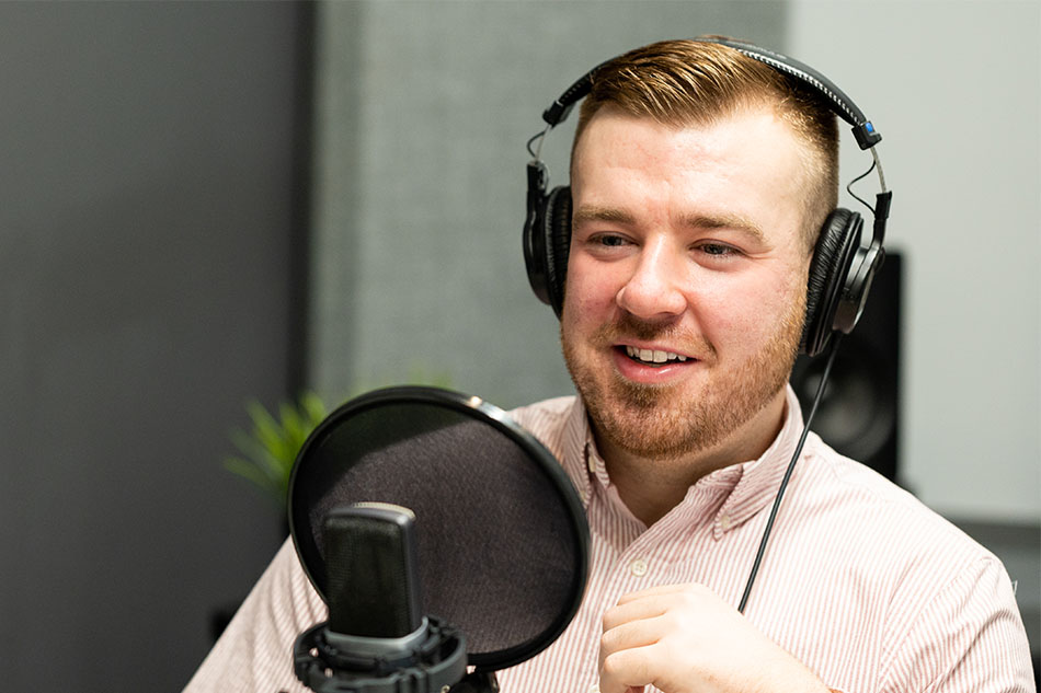 A man with a short ginger beard and a smile wears over-the-ear headphones and speaks into a microphone. 